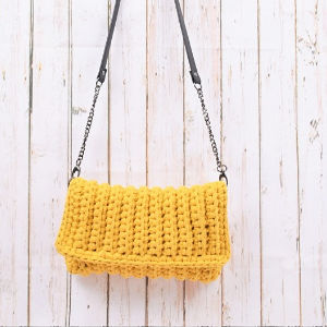 Une pochette au crochet jaune avec explication gratuite avec une lanière noire et des accents de chaîne est suspendue sur un fond de bois blanc.