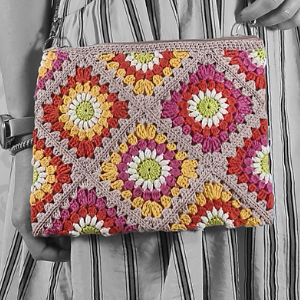 Une personne tient une pochette au crochet colorée avec explication gratuite, présentant un motif géométrique de type "granny square" en rouge, jaune, orange, vert et blanc. L'arrière-plan en niveaux de gris met en valeur une robe à rayures.