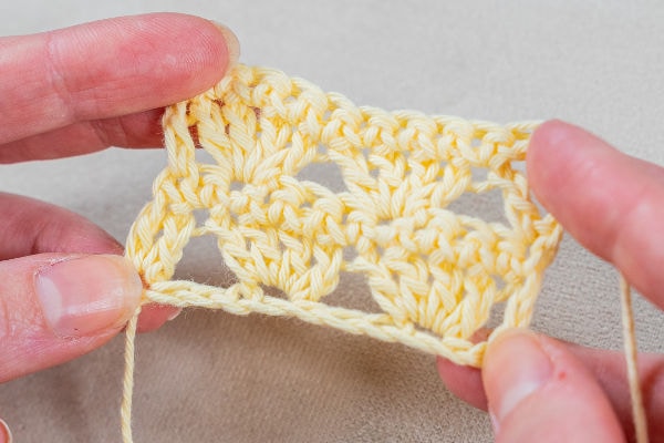 Gros plan des mains tenant un petit morceau de crochet jaune avec un motif de dentelle, montrant un point en cours avec le fil qui s'écoule.