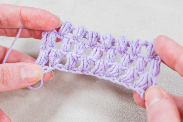 Gros plan de mains tenant un petit morceau de tissu crocheté violet clair, présentant un motif de point ouvert et en dentelle. L'arrière-plan est une surface neutre de couleur claire.