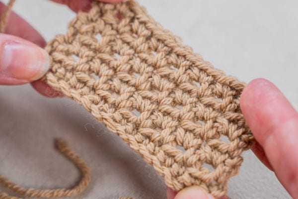 Gros plan des mains tenant un petit morceau rectangulaire de tissu crocheté marron clair, montrant clairement la texture et les points.