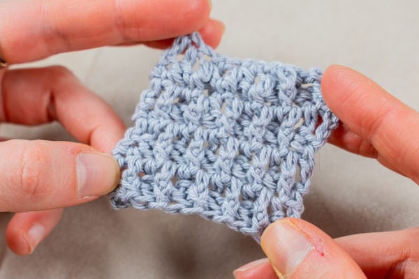 Deux mains tiennent un petit morceau carré de tissu au crochet gris clair, montrant un gros plan des points texturés d'un point au crochet original sur un fond neutre.