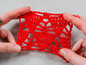 Deux mains tiennent un petit morceau rectangulaire de tissu crocheté rouge avec un motif ouvert en dentelle - un exemple de point original au crochet - sur un fond gris clair.