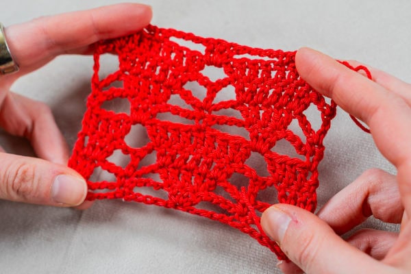 Deux mains tiennent un petit morceau de tissu rouge au crochet, présentant un motif ouvert en dentelle qui met en valeur un beau point au crochet original sur une surface claire.