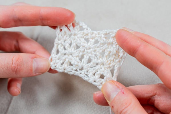 Deux mains tiennent délicatement un petit morceau de tissu blanc au crochet, réalisé selon un motif original au crochet, montrant un motif ajouré en dentelle sur un fond clair et neutre.