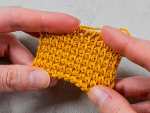 Deux mains tiennent un petit morceau de tissu jaune texturé au crochet, mettant en valeur le motif du point tunisien au crochet sur un fond neutre.