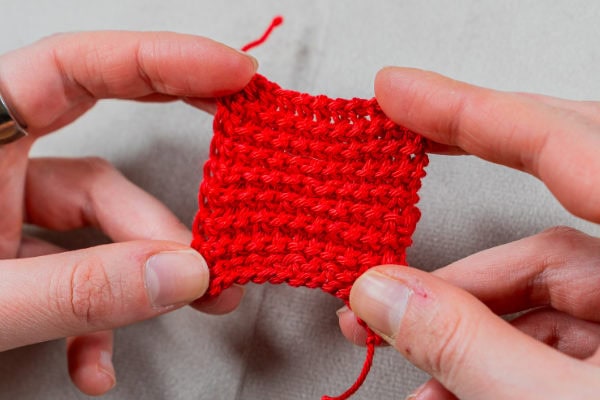 Deux mains tiennent un petit carré de tissu rouge travaillé au crochet en point tunisien sur un fond gris clair. Les points de crochet sont clairement visibles et l'on aperçoit un bout de fil détaché en haut de l'échantillon.
