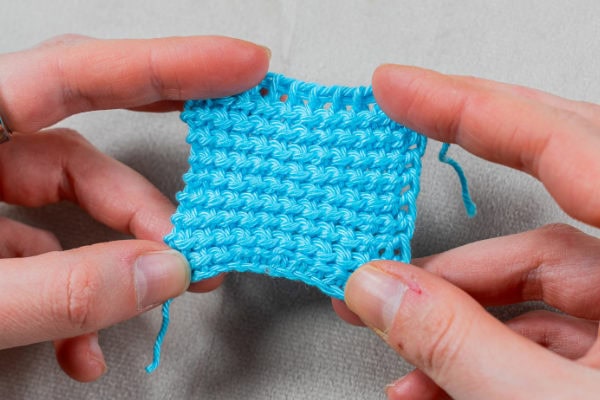 Deux mains tiennent un petit morceau carré de tissu bleu clair crocheté avec un motif texturé, mettant en valeur le point tunisien au crochet sur un fond de couleur neutre.