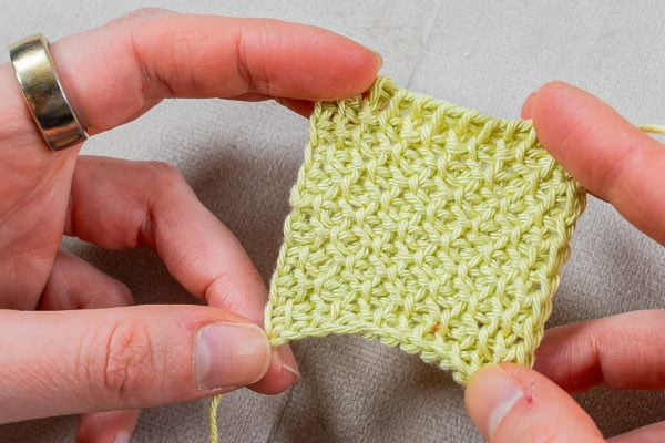 Deux mains tenant un petit carré de tissu crocheté jaune clair, présentant un motif de point texturé réalisé avec le point tunisien au crochet, sur un fond neutre.