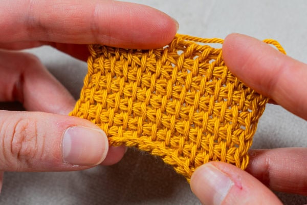 Deux mains tiennent un petit morceau de tissu jaune au crochet, montrant un motif de point texturé réalisé avec le point tunisien au crochet sur un fond gris clair.