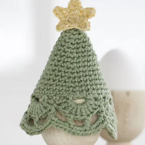 Un petit sapin de Noël vert au crochet, surmonté d'une étoile jaune, posé sur un œuf dans un coquetier en bois. L'arbre a un bord décoratif festonné près du bas.