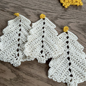 Trois décorations de sapin de Noël au crochet, blanches avec un sommet jaune, sont disposées côte à côte sur une surface en bois. Une petite pièce de crochet jaune est partiellement visible à l'arrière-plan.