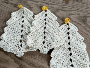 Trois pièces en forme d'arbre crochetées en fil blanc cassé avec de petites pointes jaunes, disposées côte à côte sur une surface en bois, inspirées d'un diagramme sapin de noël au crochet.