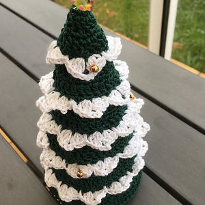 Un petit sapin de Noël crocheté à couches vertes et blanches, décoré de minuscules perles dorées, est posé sur une surface en bois gris.