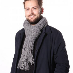 Un homme barbu sourit, vêtu d'une écharpe grise épaisse homme au crochet et d'un manteau sombre, sur un fond blanc uni.