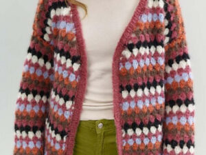 Une personne portant un confortable gilet en crochet pour femme, ouvert sur le devant, avec un motif coloré et épais en rose, violet, noir, blanc et orange, associé à une chemise beige clair et un pantalon vert olive.