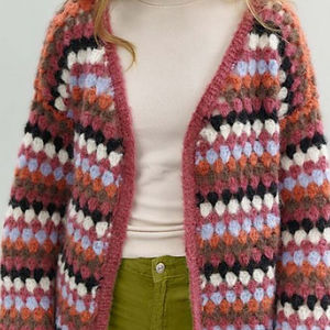 Une personne porte un gilet coloré en crochet pour femme avec un motif géométrique en rose, violet, noir, blanc et orange sur un haut beige clair et un pantalon vert olive. La tête de la personne n'est pas entièrement visible.