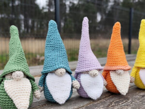 Cinq lutins au crochet colorés avec de longs chapeaux et des barbes blanches sont assis en rang sur un banc en bois, en plein air, avec une clôture floue et des arbres en arrière-plan.