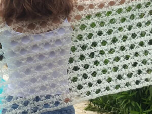 Une personne vêtue d'un haut blanc et d'un jean bleu brandit un grand châle aéré fait à la main au crochet en fil blanc, mettant en valeur son motif ouvert et dentelé en plein air dans un jardin - un parfait Modèle d'Étole au Crochet Gratuit.