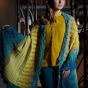 Une femme portant un chemisier jaune et un pantalon sarcelle est drapée dans un grand châle multicolore crocheté avec des nuances dégradées de bleu, de vert et de jaune. Elle se tient à l'intérieur, près d'un ouvrage métallique orné.