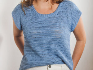 Une personne portant un haut bleu clair, à manches courtes, en crochet - parfait comme modèle de haut pour femme au crochet gratuit - et un pantalon blanc se tient debout sur un fond blanc uni.