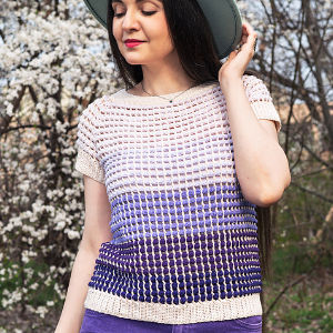 Une femme portant un haut en crochet dégradé bleu et violet et un chapeau à large bord se tient à l'extérieur, près d'arbres en fleurs, touchant doucement son chapeau et souriant les yeux fermés.