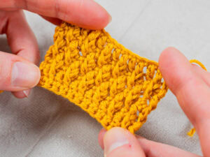 Deux mains tiennent un petit morceau de tissu crocheté jaune vif, montrant des points détaillés réalisés au crochet alpin. L'arrière-plan est une surface légère et texturée.