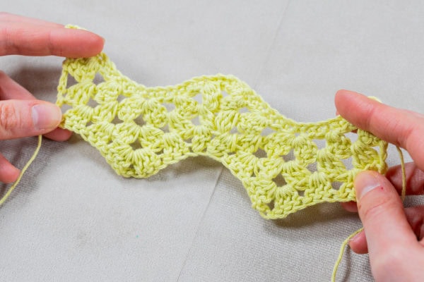 Deux mains tiennent un morceau de tissu crocheté jaune, présentant un motif de crochet granny au à points ondulés et festonnés, sur un fond gris clair.