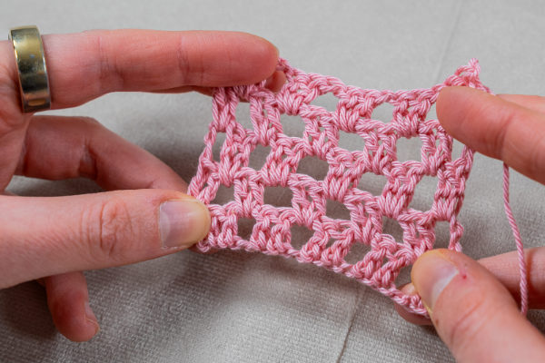 Deux mains tiennent un petit morceau de tissu rose au crochet avec un motif carré et ajouré, mettant en valeur les points classiques de granny au crochet sur un fond gris clair.