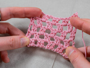 Gros plan de deux mains tenant un petit morceau de tissu crocheté rose en treillis ouvert sur un fond gris clair. Le crochet présente un motif carré, en forme de grille, qui ressemble à la technique du crochet granny au point.