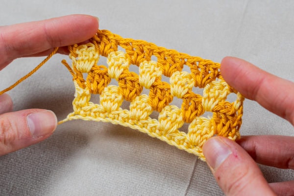 Gros plan sur des mains tenant un petit échantillon réalisé au crochet point granny, avec des rangées alternées de fils jaune et orange, sur un fond de tissu clair.