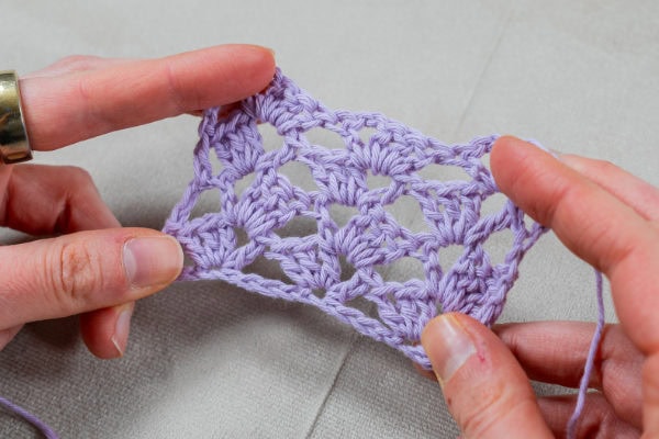 Deux mains tiennent un petit morceau rectangulaire de dentelle au crochet violet clair avec un motif au crochet granny au à pointe triangulaire ouverte, sur un fond de tissu gris clair.