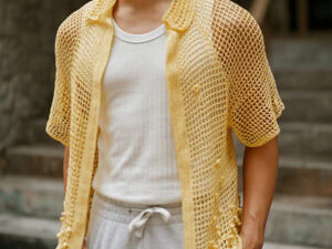 Une personne portant un T-shirt blanc côtelé, un pantalon gris clair à cordon de serrage et un gilet jaune ouvert à manches courtes "pull d'été au crochet facile", debout en plein air près de marches en pierre.