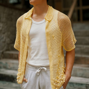 Une personne portant une chemise en crochet jaune à manches courtes sur un débardeur blanc côtelé et un pantalon de survêtement gris clair, debout à l'extérieur près de marches en pierre.