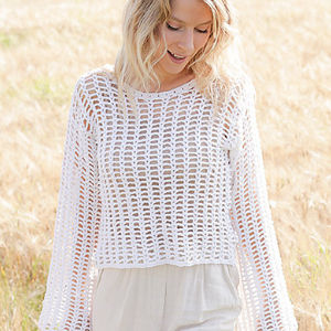Une femme aux cheveux blonds ondulés se tient à l'extérieur, vêtue d'un ample pull en crochet blanc à manches longues sur un haut et un pantalon de couleur claire. Elle regarde vers le bas et sourit dans un champ ensoleillé.