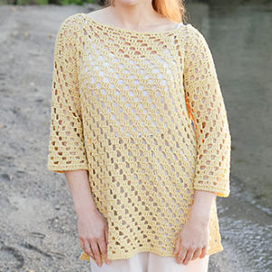 Une personne se tenant à l'extérieur, au bord de l'eau, portant un pull-over jaune crocheté à manches trois-quarts sur un haut blanc et un pantalon de couleur claire. Le pull présente un motif ouvert en dentelle.