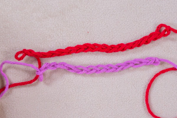 Deux fils, l'un rouge et l'autre violet, sont crochetés en chaînettes simples et posés parallèlement sur une surface texturée beige clair. Les extrémités de chaque fil sont visibles.