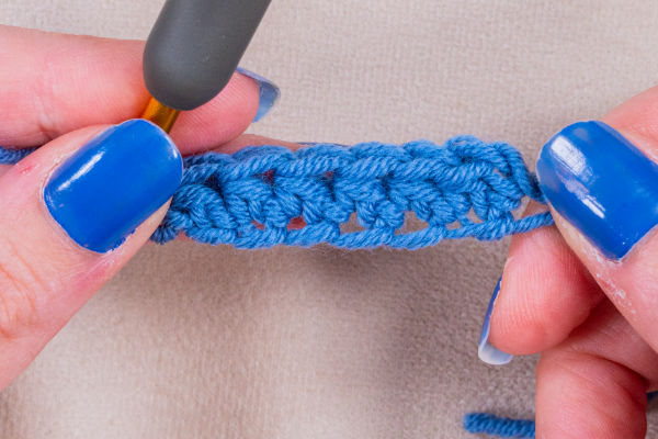 Gros plan sur des mains recouvertes de vernis à ongles bleu tenant un crochet, travaillant habilement un fil bleu pour former une chaînette en crochet, formant des points nets sur un fond de tissu clair.