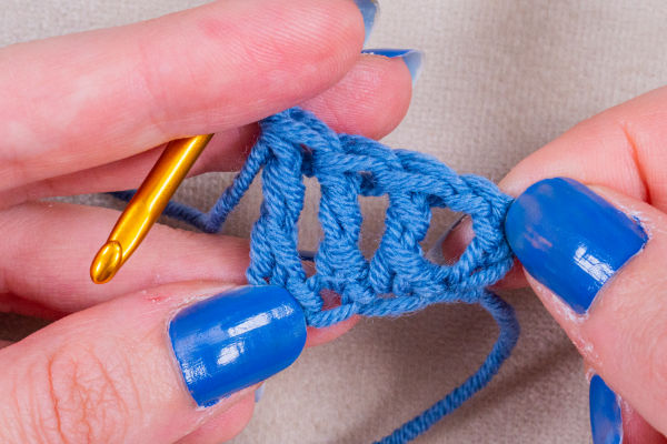 Gros plan sur des mains recouvertes de vernis à ongles bleu tenant un fil bleu et un crochet doré, travaillant sur un projet de chaînette en crochet avec plusieurs boucles cousues visibles.