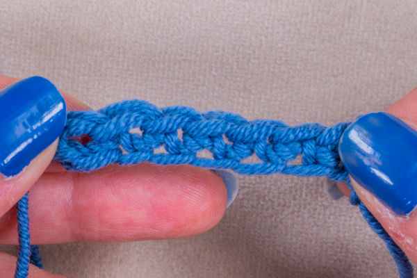 Gros plan de mains aux ongles peints en bleu tenant une rangée de chaînette en crochet bleue sur fond beige.