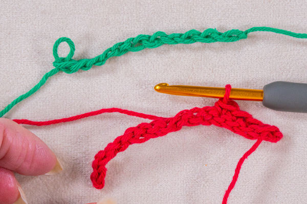 Gros plan d'un projet de crochet montrant un crochet doré travaillant avec du fil rouge et une chaînette en crochet verte terminée au-dessus, tous deux sur un fond de tissu clair. Une main tient l'extrémité du fil rouge.