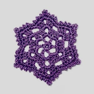 Un hexagone violet crocheté avec un motif complexe en dentelle est posé sur un fond gris clair uni. Le fil forme des espaces ouverts et des détails texturés à l'intérieur de l'étoile à six branches.