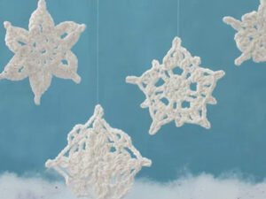 Quatre flocons de neige au crochet blancs sont suspendus par des ficelles presque invisibles sur un fond bleu, avec une couche de matière blanche duveteuse ressemblant à de la neige au fond.