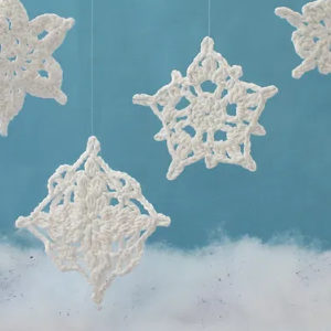 Des flocons de neige blancs au crochet sont suspendus par des fils clairs sur un fond bleu, avec un tissu blanc duveteux au fond, ressemblant à des nuages ou à de la neige.