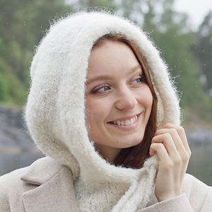 Une femme sourit en remontant la capuche de son pull blanc duveteux. Elle est à l'extérieur, avec des arbres et de l'eau en arrière-plan.