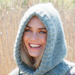 Une personne souriante aux yeux bleus et aux cheveux blonds porte un épais pull tricoté gris clair à capuche à l'extérieur, avec de hautes herbes beiges en arrière-plan flou.