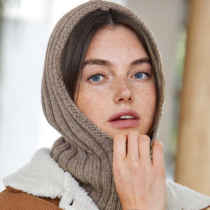 Une femme aux yeux bleus et aux taches de rousseur porte une capuche beige tricotée et une veste en shearling beige. Elle regarde légèrement sur le côté, sa main touchant son menton.