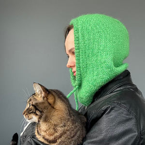 Une personne vêtue d'une capuche en tricot vert vif et d'une veste noire tient un chat tabby, tous deux tournés vers la gauche sur un fond gris uni.