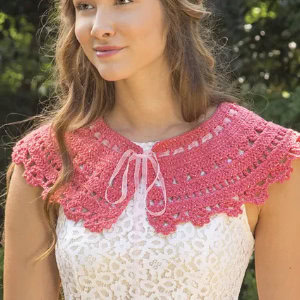 Une femme portant une robe en dentelle blanche et un col au crochet rose avec explication, aux bords festonnés et noué par un ruban à l'encolure, se tient à l'extérieur avec de la verdure en arrière-plan.