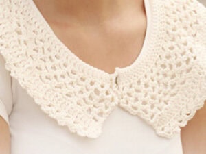 Gros plan d'une personne portant un col au crochet de couleur crème avec explication, superposé à une chemise blanche. Le col en dentelle crochetée présente un délicat motif ajouré et se ferme au centre du devant.